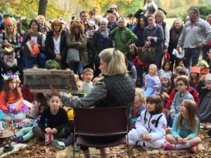 Children and families gather around as someone reads a book in the park.