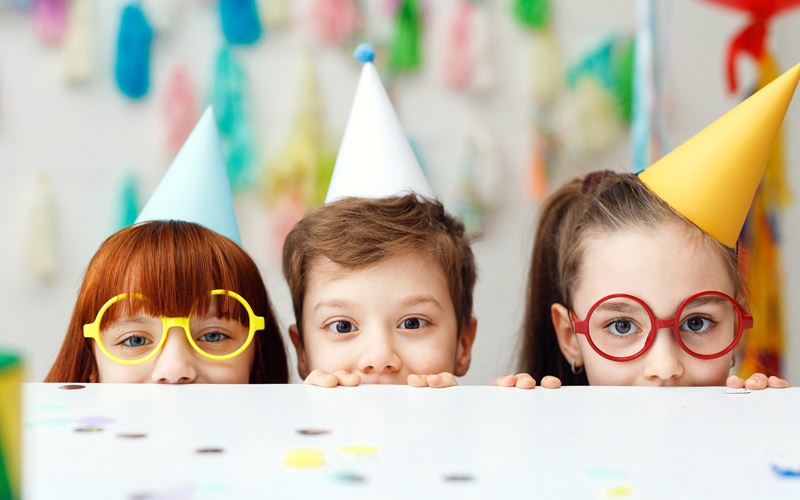 Three kids peak over a table top wearing birthday hats.