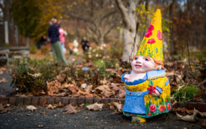 Sylvia the gnome sits in the Nadeau Garden with her yellow floral hat and dress. Behind her, visitors wander among the other gnomes.
