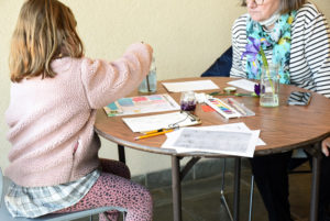 A young girl sits at a table with her grandmother during an April Vacation week watercolor class.