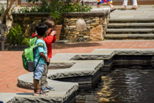 Two children stand with their discovery backpacks looking into the water of the turtle fountain. Drop-in activities.