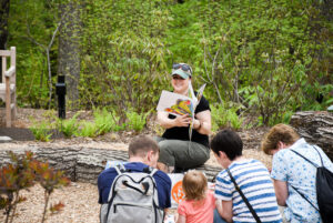 Families and children sit in The Ramble and listen to a story.
