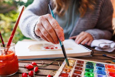 a person's hand dipping into a watercolor paint palette with a painting pad in front of them
