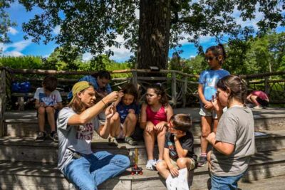 Students from age 5-12, gather around on the stairs under a tree with an instructor giving a nature lesson