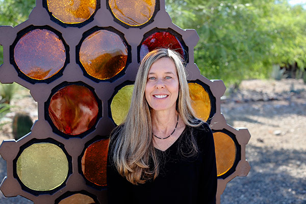 Artist, Alex Heveri, smiling in front of her glass sculpture made of multiple warm-toned glass circles