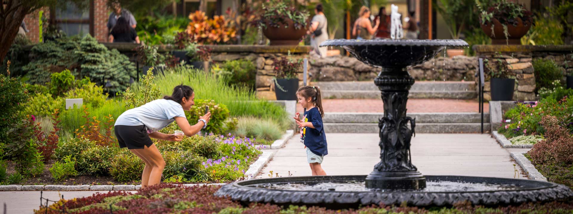 A guardian taking a picture of an excited child next to the water fountain in the Garden of Inspiration during a summer evening