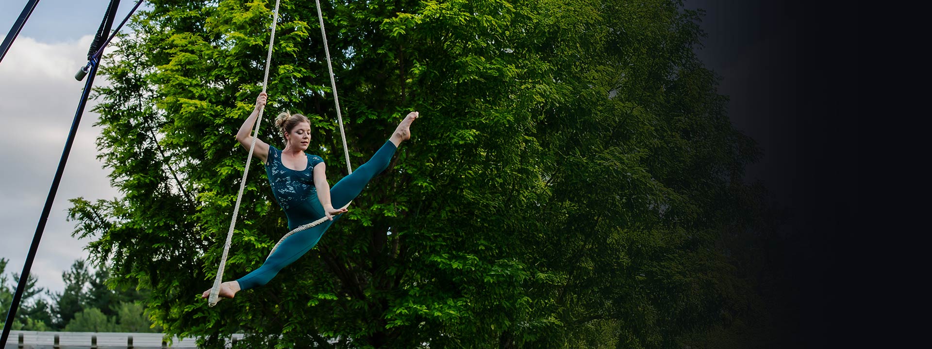 An aerialist hanging from ropes doing a ballet pose, outdoors with a tree behind her