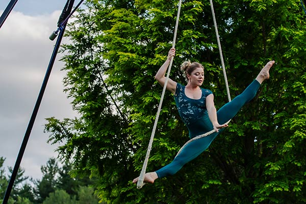 An aerialist hanging from ropes doing a ballet pose, outdoors with a tree behind her