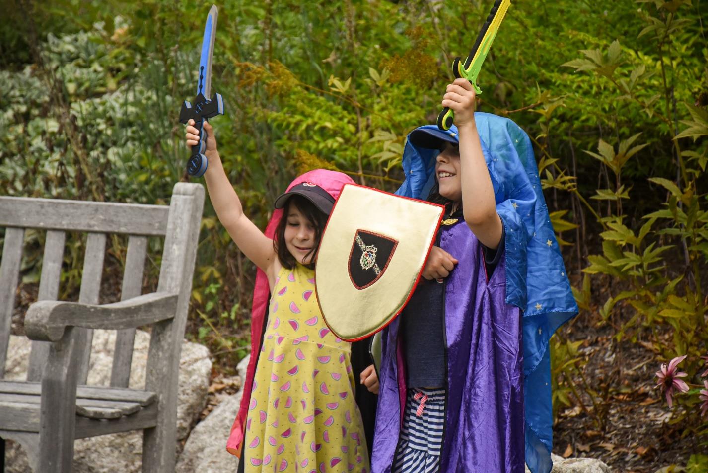 Two kids dressed up colorfully wearing blankets as capes, and a toy sword and shield in their hands