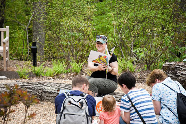 An instructor reading to a group of youth in parents in a nature setting, sitting on logs