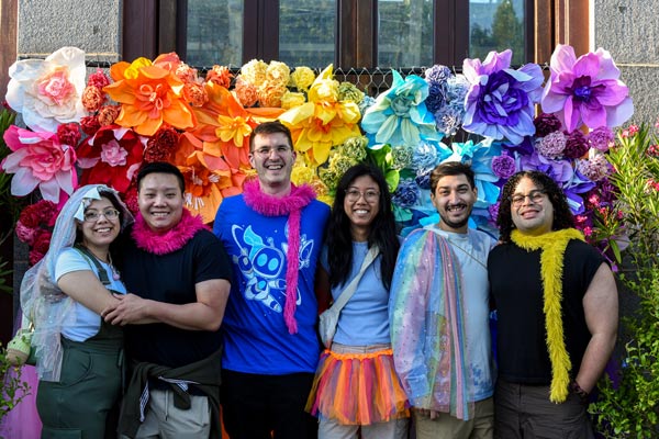 Dazzling group of friends in Pride accessories pose in front of a background made of rainbow craft flowers