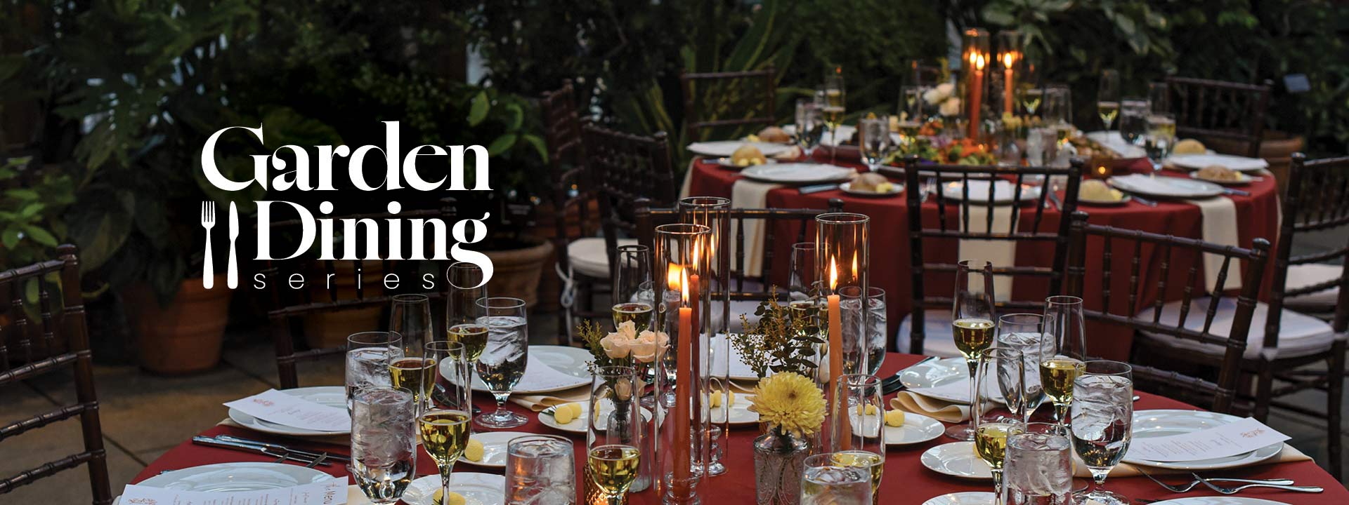 Table set up elegantly with candles and silverware on a red table cloth. Dining is located in the conservatory.